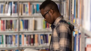 شاب يرتدي قميصاً مزخرفاً يقف بجانب رفوف الكتب في مكتبة، يقرأ كتاباً بتركيز.