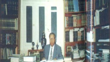 رجل يجلس خلف مكتب في مكتبة تحتوي على رفوف مليئة بالكتب. يحيط به أكوام من الكتب والمجلدات، مع نافذة خلفه. الأجواء تعكس حب المعرفة والثقافة.