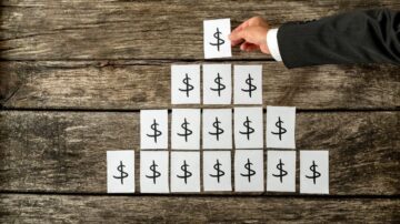 رجل يرتدي بدلة يضع ورقة تحمل علامة الدولار على هيكل هرمي من أوراق تحمل نفس الرمز، على سطح خشبي داكن. الصورة تعكس مفهوم المال والاستثمار.