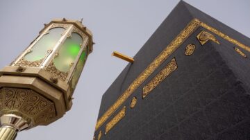 صورة توضح الكعبة المشرفة من زاوية مرتفعة، مع وجود فانوس مزخرف بجانبها. الكعبة مغطاة بكسوة سوداء مزينة بنقوش ذهبية، مما يبرز جمالها وروحانيتها.