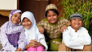 مجموعة من الأطفال يرتدون ملابس تقليدية، يجلسون معًا مبتسمين في بيئة خارجية. يظهر في الصورة طفلان وطفلتان، واحدة منهن ترتدي حجابًا. تعكس الصورة الفرح والترابط بين الأصدقاء.