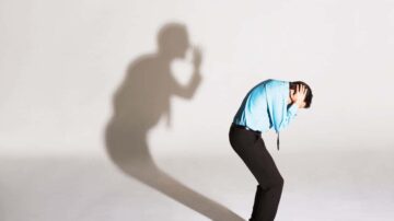 رجل يرتدي قميصًا أزرق ويضع يديه على رأسه في وضعية انكماش، بينما تظهر ظلاله خلفه بشكل درامي على الحائط. تعبر الصورة عن مشاعر القلق أو الضغط.