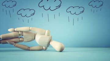 دميّة خشبية مستلقية على سطح أزرق، مع سحب مرسومة تتساقط منها أمطار. تعكس الصورة شعور الحزن أو الاستسلام.