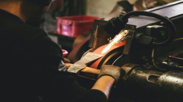 شخص يعمل في ورشة حرفية، يستخدم آلة لطحن المعدن، تتطاير الشرر من قطعة المعدن أثناء المعالجة، خلفية تحتوي على أدوات وأماكن عمل مختلفة.