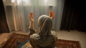 امرأة ترتدي حجابًا، ترفع يديها في دعاء، تجلس على سجادة صلاة في غرفة مضيئة بأشعة الشمس، مع ستائر شفافة وخلفية داكنة.