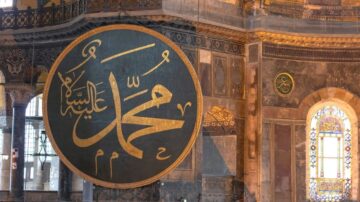خط كبير باللون الذهبي يحمل اسم محمد عليه السلام في قبة آيا صوفيا، محاط بتفاصيل معمارية رائعة ونوافذ ملونة.