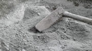 صورة تظهر مجرفة مغمورة في كومة من الرمل أو الأسمنت الرمادي، مع تفاصيل دقيقة للمواد الجافة. تبرز الصورة القوام الناعم والترابي للسمنت، مما يعكس استخدامه في أعمال البناء والتشييد.