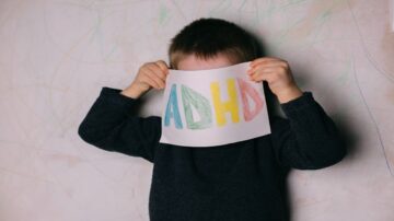 طفل صغير يحمل ورقة مكتوب عليها "ADHD" بألوان زاهية، يغطي وجهه بها، مع خلفية جدارية مغطاة بخطوط ملونة.