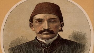 صورة ملونة لرجل يرتدي طربوشًا أحمر وقميصًا داكنًا، يظهر بملامح وجه واضحة، بما في ذلك شارب كثيف ونظرة جدية. الخلفية بلون بيج فاتح، مما يبرز تفاصيل الصورة التاريخية.