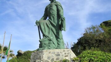 تمثال كبير لشخصية تاريخية تحمل سيفًا، يقف على قاعدة صخرية. يتميز التمثال بتفاصيل دقيقة وملامح قوية، محاط بالنباتات الخضراء وأشجار النخيل في الخلفية. السماء زرقاء مع بعض الغيوم.