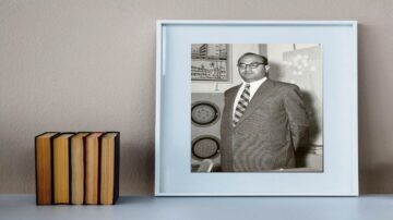 صورة بالأبيض والأسود لرجل يرتدي بدلة رسمية ونظارات، يقف بجانب إطار صورة في غرفة ذات جدران بلون محايد. يوجد على الطاولة مجموعة من الكتب الملونة.