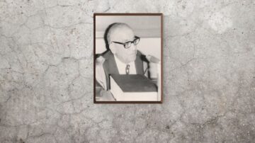 صورة بالأبيض والأسود لرجل مسن يرتدي نظارات وقميصًا رسميًا، يجلس أمام كتاب على طاولة. الإطار خشبي ويظهر على خلفية جدارية رمادية.