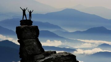 شخصان يحتفلان بقمّة جبلية، silhouetted against سلسلة جبال ضبابية في خلفية. السماء تميل إلى الأزرق مع سحب خفيفة، مما يخلق مشهدًا مدهشًا للطبيعة.