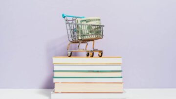 عربة تسوق صغيرة تحتوي على نقود موضوعة على مجموعة من الكتب مع خلفية بنفسجية. تعبر الصورة عن مفهوم التعلم والاستثمار المالي.