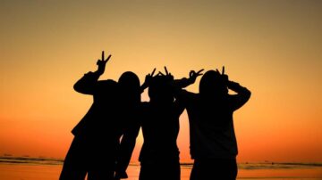 ظلال ثلاث أشخاص يقفون معًا على الشاطئ أثناء غروب الشمس، يرفعون أصابعهم في إشارة النصر. السماء تتدرج بين الألوان البرتقالية والوردية، مما يخلق مشهدًا جميلًا ومليئًا بالطاقة الإيجابية.