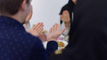 مجموعة من الأشخاص يجلسون حول طاولة، مع التركيز على يدي شخصين يصفقون. يتواجد شخص يرتدي زيًا تقليديًا أسود وآخر بقميص مزخرف بأزرار. تعكس الصورة أجواء من التعاون والمشاركة في احتفال أو مناسبة اجتماعية.