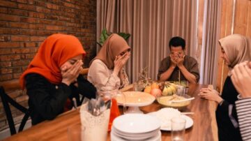 مجموعة من الأشخاص يجلسون حول طاولة طعام، مع تغطية وجوههم بأيديهم. يتكون المشهد من أربعة أشخاص يرتدون ملابس تقليدية، ويبدو أنهم في لحظة تأمل أو دعاء. توجد أطباق من الطعام والفواكه على الطاولة، مع خلفية من جدران من الطوب والأقمشة.