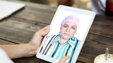 امرأة ترتدي حجابًا ورديًا، تنظر إلى شاشة جهاز لوحي بينما تحملها بيدها. تظهر خلفية طبيعية مع طاولة خشبية ومشروبات.