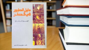 كتاب دليل الطيور في قطر، يغطي أنواع الطيور المختلفة في المنطقة، مصحوبًا بصور ملونة. يظهر الكتاب على طاولة خشبية بجانب مجموعة من الكتب.