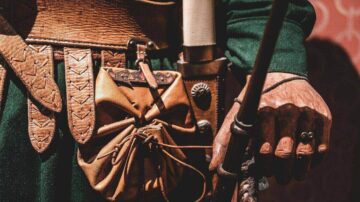 يد تظهر تفاصيل ملابس تاريخية، مع حزام جلدي وقبعة صغيرة. اليد ممسكة بعصا، وتحتوي على حقيبة جلدية مربوطة بحبل. الخلفية تحتوي على نمط مزخرف، مما يضيف عمقًا للصورة.