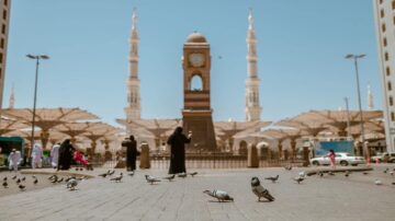 مشهد من الحرم المدني، يظهر فيه مبنى الساعة الشهير مع مآذن مرتفعة، بينما تتجول الطيور في الساحة. يرتدي بعض الأشخاص ملابس تقليدية، مما يضفي طابعًا ثقافيًا على الصورة. السماء صافية والشمس مشرقة، مما يبرز جمال المكان.