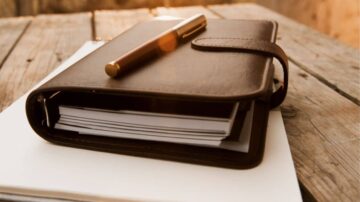 مفكرة جلدية بنية اللون مع قلم على سطح خشبي، بجانب أوراق بيضاء. الضوء الطبيعي ينعكس على الجزء العلوي، مما يضفي جواً دافئاً ومريحاً.