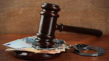 صورة تضم مطرقة قانونية مصنوعة من الخشب، موضوعة على مجموعة من الأوراق النقدية الأوروبية، مع قيود معدنية بجانبها. تعكس الصورة تداخل المال مع القوانين والعدالة.