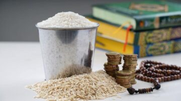 وعاء معدني مملوء بالأرز الأبيض مع حبوب الأرز البني بجانبه، وترتيب من العملات المعدنية، وخلفية تحتوي على كتب دينية ملونة.