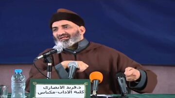 دوريد الأنصاري يتحدث في مؤتمر، مرتديًا عمامة، ويشير بيده خلال إلقاء محاضرة، مع وجود ميكروفونات وزجاجة ماء على الطاولة.