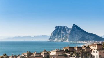 منظر خلاب لجبل جبل طارق يطل على البحر، مع منازل تقليدية في المقدمة. السماء زرقاء وصافية، والمياه تتلألأ تحت أشعة الشمس.