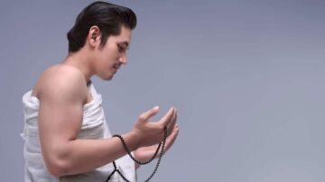 رجل يحمل سبحة في يديه، يبدو في حالة تأمل. يرتدي منشفة بيضاء ويظهر جانب وجهه في وضع جانبي، مع خلفية رمادية بسيطة. تعكس الصورة الهدوء والتفكير العميق.