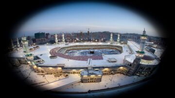 صورة جوية للحرم المكي، تظهر الكعبة المشرفة وسط الحرم، محاطة بالمصلين خلال وقت الصلاة. الأضواء تضيء المكان، مع وجود معالم البناء الحديثة حوله.