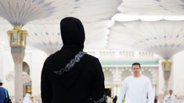 امرأة ترتدي حجابًا أسود وتواجه سماءً مغطاة بأقواس كبيرة، بينما يسير رجل في ملابس بيضاء في الخلفية. تعكس الصورة أجواء المدينة المنورة، مع التركيز على العمارة الإسلامية الجميلة.