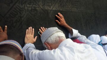 أشخاص يلمسون الكعبة أثناء الصلاة في مكة، مع التركيز على الأيادي والملابس التقليدية. الخلفية تظهر النقوش الإسلامية على الكعبة.