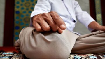 يد شخص جالس في وضعية التأمل، مع راحة اليد مستندة على الركبة، في بيئة هادئة مزينة بنقوش تقليدية.