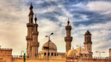 مسجد تاريخي بأبراج عالية وقبة كبيرة، يبرز جمال العمارة الإسلامية تحت سماء زرقاء ملبدة بالغيوم. يحيط بالمسجد أعمدة معمارية مميزة، مما يعكس التراث الثقافي الغني.