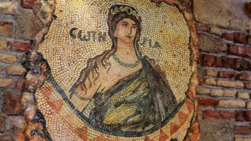 فسيفساء قديمة تظهر امرأة ترتدي لباسًا تقليديًا، محاطة بحدود مزخرفة. التفاصيل الدقيقة تبرز في الألوان الغنية والخطوط، مما يعكس التراث الثقافي والفني. الجدران الحجرية المحيطة تضيف عمقًا للمشهد.