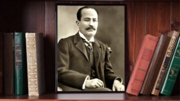 صورة تاريخية لرجل يرتدي بدلة رسمية مع ربطة عنق، يجلس على كرسي. الصورة مؤطرة وتعرض على رف مليء بالكتب القديمة، مما يضفي طابعاً ثقافياً وأدبياً على المشهد.