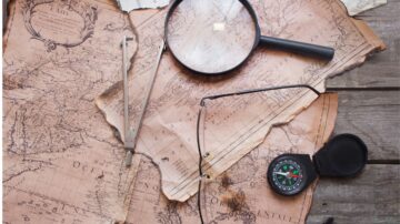 خريطة قديمة تظهر تفاصيل جغرافية مع عدسة مكبرة، وأداة قياس، وبوصلة. العناصر موضوعة على سطح خشبي، مما يضيف جواً تاريخياً واستكشافياً.