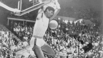 لاعب كرة سلة من جامعة كاليفورنيا في لوس أنجلوس (يو سي إل إيه) يقوم بعملية دنك مثيرة تحت سلة، مع وجود لاعبين آخرين من فريق ستانفورد في الخلفية. الملعب مليء بالجماهير.