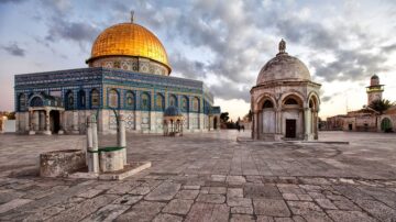 قبة الصخرة في القدس تظهر تحت سماء غائمة، مع قبة ذهبية لامعة. المباني المحيطة تتميز بتفاصيل معمارية إسلامية تقليدية، مع حجر رملي وألوان زاهية. الفناء الواسع يضم أعمدة ومصلى صغير، مما يبرز جمال الموقع الديني والتاريخي.