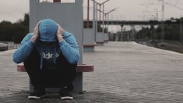 شاب يجلس على رصيف محطة قطار، مرتديًا سترة زرقاء مع غطاء رأس، يمسك برأسه بيديه، ويظهر عليه الإحباط. خلفه، توجد أعمدة السكك الحديدية وأجواء قاتمة تعكس شعور الوحدة.