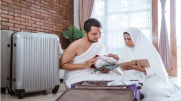 رجل وامرأة يجلسان على الأرض بجانب حقيبتين سفر مفتوحتين، يراجعان الملابس والملاحظات. الرجل يرتدي الإحرام والمرأة ترتدي الحجاب، في غرفة ذات جدران من الطوب وأرضية مريحة.