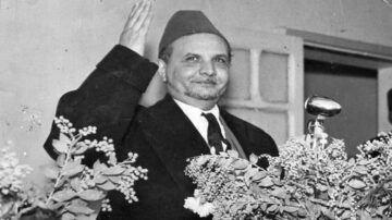 رجل يرتدي عمامة وخطاب رسمي، يلوح بيده وسط مجموعة من الزهور، خلال حدث أو خطاب رسمي.