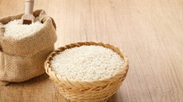 أرز أبيض في وعاء مصنوع من القش مع كيس من الأرز بجانبه. خلفية خشبية تعزز من مظهر الصورة.