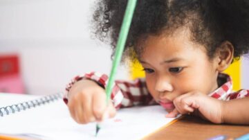 طفل صغير ذو شعر مجعد، يرتدي قميصًا مخططًا، ينشغل بالرسم في دفتر. يظهر التركيز على وجهه بينما يمسك قلمًا ملونًا.