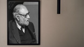 صورة بالأبيض والأسود لرجل مسن ذو لحية وشعر رمادي، يرتدي نظارات وقميص أسود مع سترة. يظهر الرجل في ملف شخصي، مع إطار صورة أسود على حائط بلون محايد.