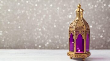 فانوس معدني ذهبي مزخرف مع زجاج بنفسجي، موضوع على سطح خشبي أبيض مع خلفية لامعة.