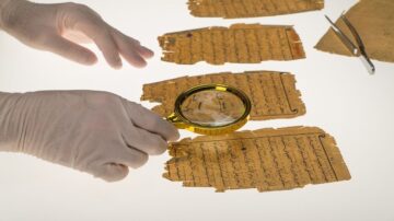 يد تظهر تحتها أوراق تاريخية قديمة مع نصوص مكتوبة بخط عربي. يتم استخدام عدسة مكبرة لفحص التفاصيل الدقيقة للكتابات. أدوات مساعدة مثل الملقط موجودة بجانب الأوراق.