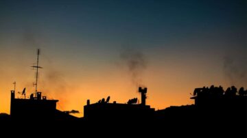 غروب الشمس خلف أسطح المباني، مع ظلال للأبراج والأطباق الهوائية. السماء تتدرج من الأزرق إلى البرتقالي، مع بعض الدخان يتصاعد.
